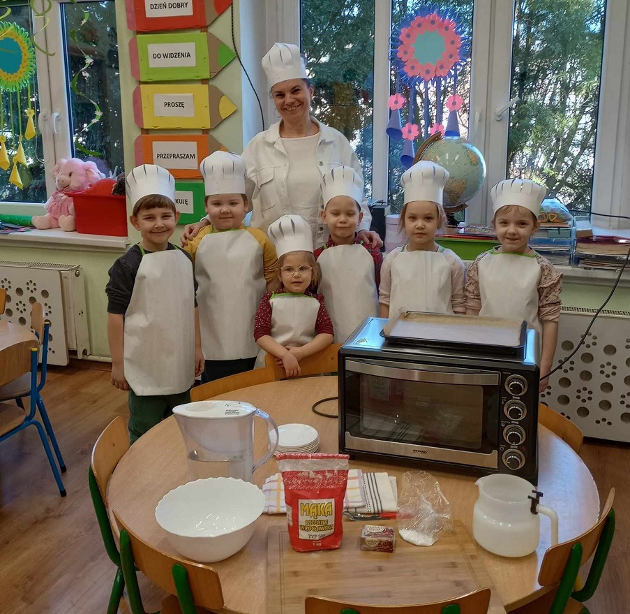 Dzieci w czapkach i fartuszkach kucharza, na stole piekarnik, mąka i naczynia